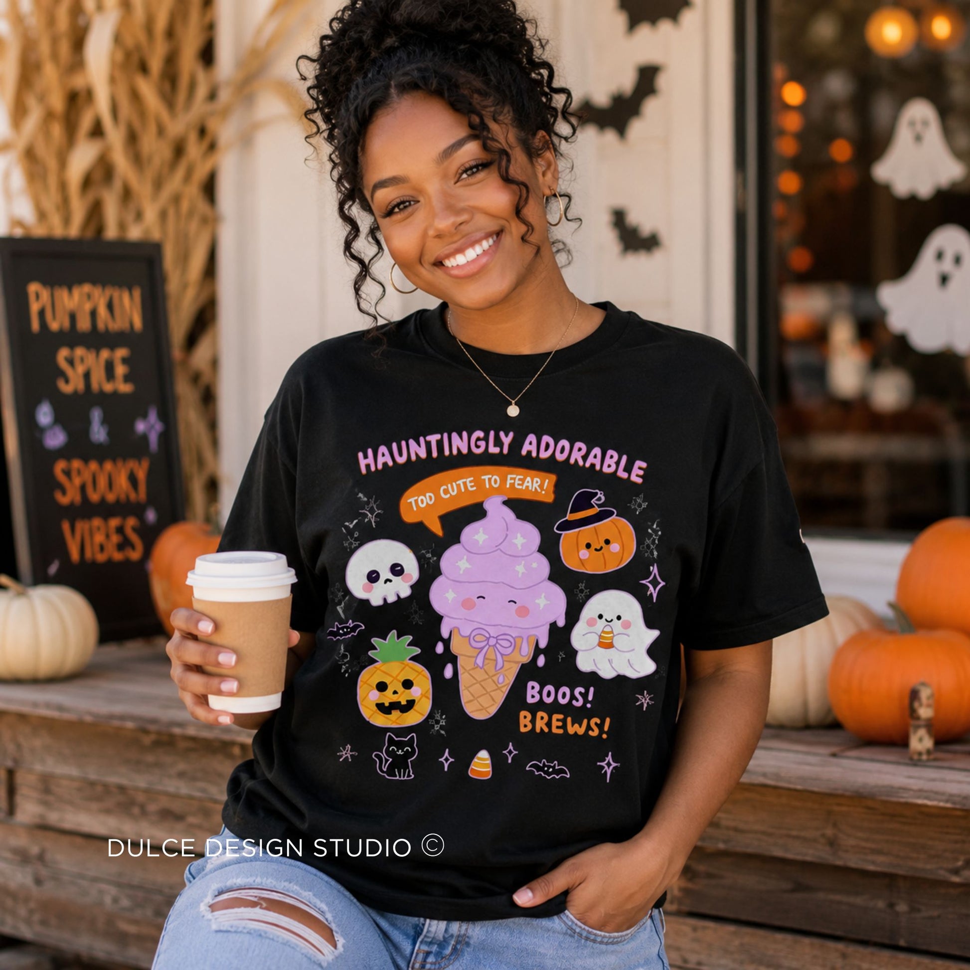 A woman wearing a black graphic t-shirt with a cute Halloween ice cream design, holding a coffee cup in a fall-themed storefront setting.