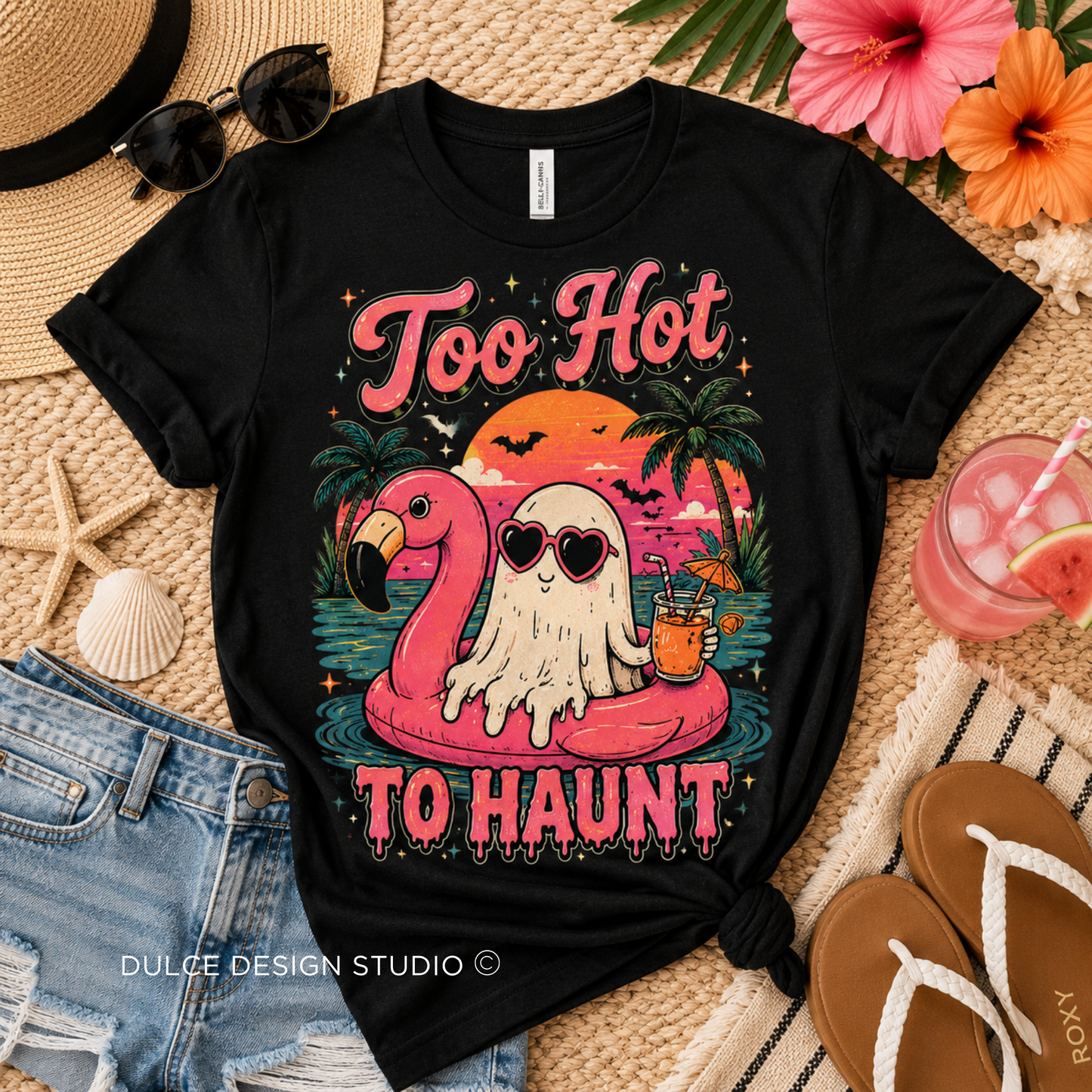 A black graphic t-shirt laid flat on a woven straw mat with beach-themed props including sunglasses, a straw hat, denim shorts, flip-flops, flowers, and a drink. The shirt features a colorful graphic of a ghost in sunglasses sitting on a pink flamingo float with the text 'Too Hot TO HAUNT'.