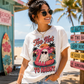 A woman wearing a white graphic t-shirt with a pink flamingo and 'Too Hot To Haunt' design, standing on a sunny beach with palm trees and colorful signs in the background.