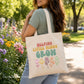 A woman outdoors carrying a cream canvas tote bag with the text 'HELPING LITTLE MINDS GROW' and colorful flower illustrations. The background shows a blurred park scene with flowers and trees.