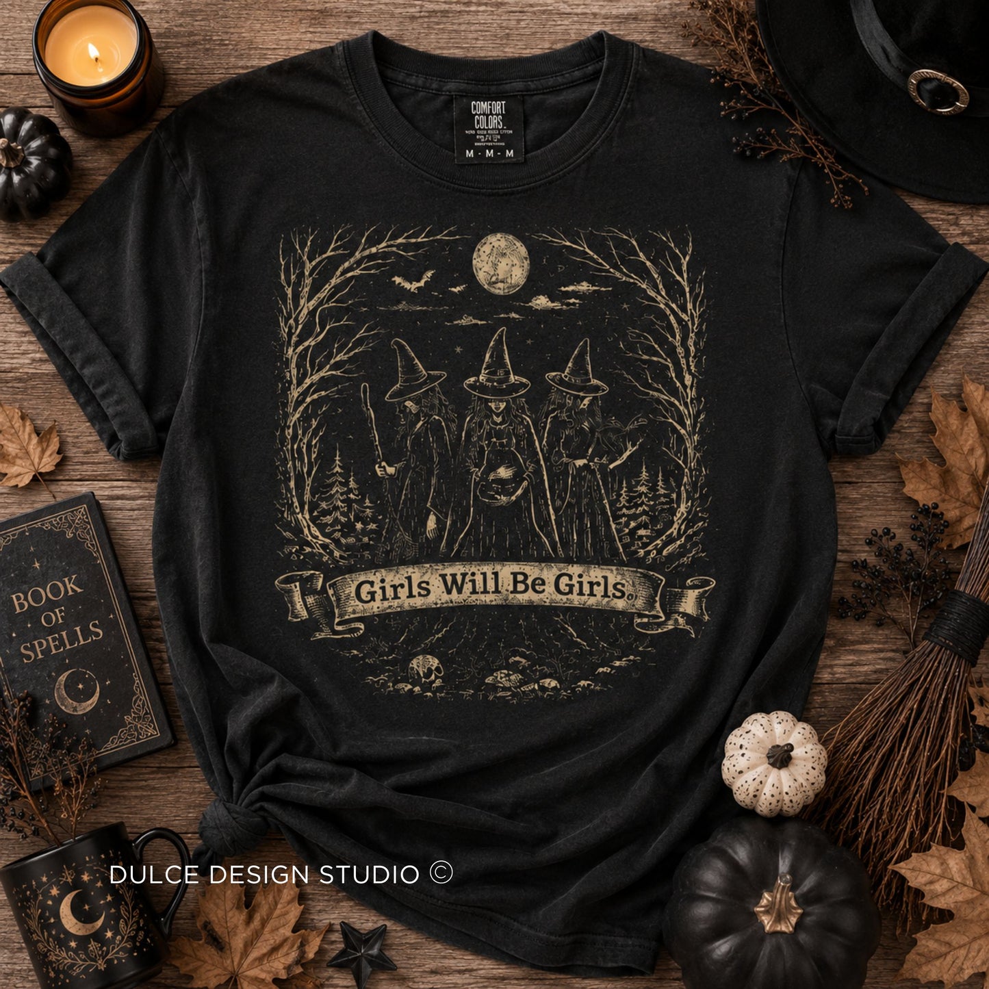 A black graphic t-shirt featuring a vintage-style illustration of three witches with hats under a full moon, with the text 'Girls Will Be Girls' on a banner. The shirt is displayed in a flat-lay arrangement with autumn-themed props including a candle, a book of spells, a witch's hat, and small pumpkins on a rustic wooden background.