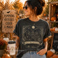A woman wearing a dark charcoal gray graphic t-shirt with a witch illustration and the text 'Girls Will Be Girls', standing in a pumpkin patch setting.