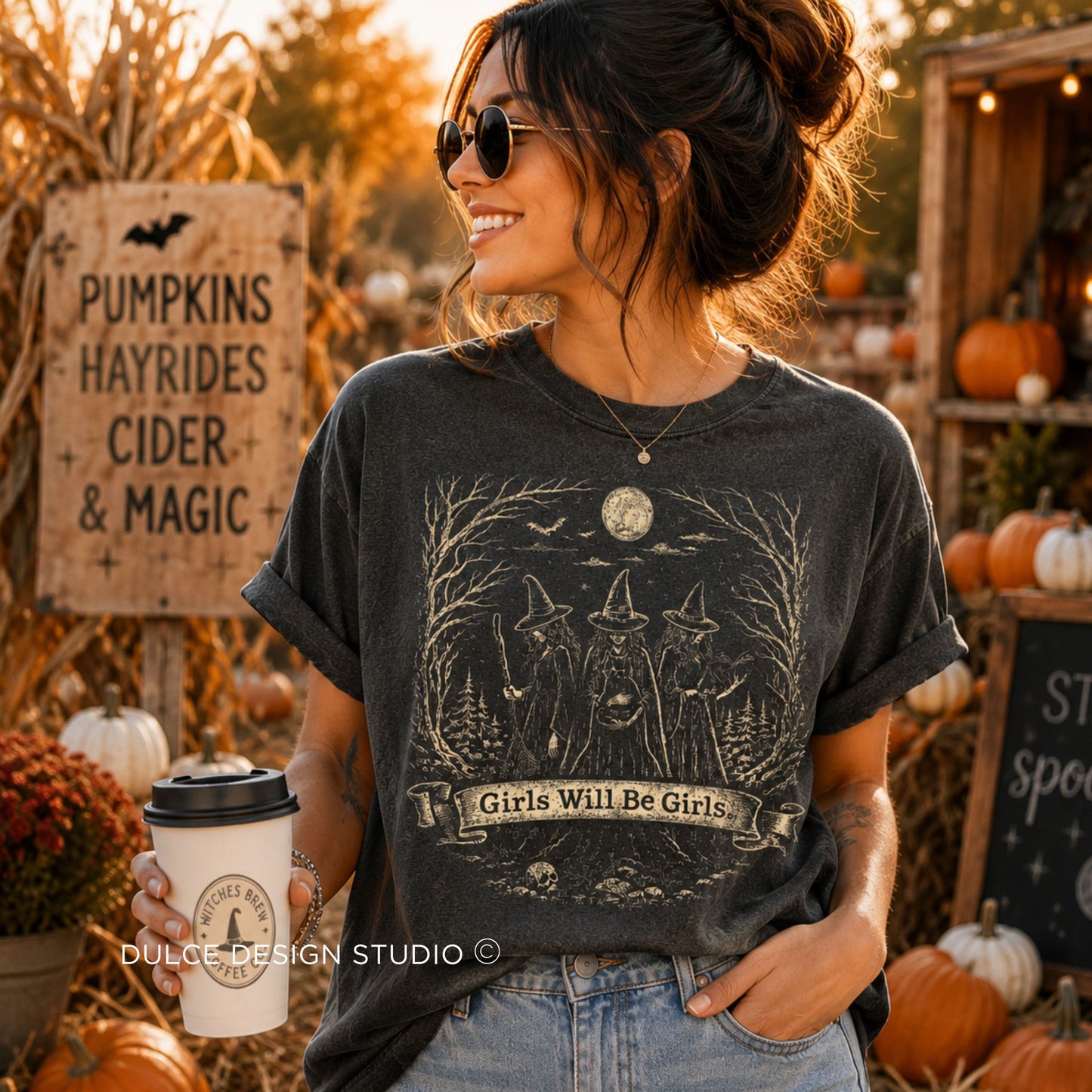 A woman wearing a dark charcoal gray graphic t-shirt with a witch illustration and the text 'Girls Will Be Girls', standing in a pumpkin patch setting.