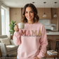 Woman wearing a pink sweatshirt with 'MAMA' text, holding a mug in a kitchen.