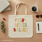Beige tote bag with 'YOUNG GIFTED & BLACK' text on a wooden surface with coffee and stationery items.